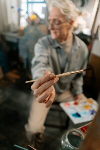 Elderly painter in art studio extending a paintbrush towards the viewer with an artistic blurred background.